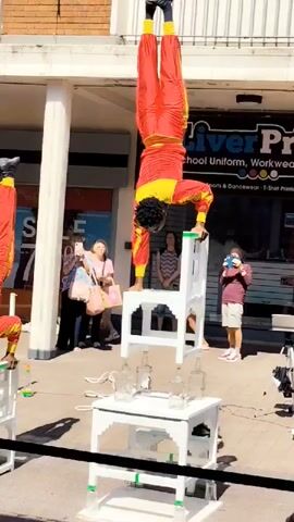 A male performer in a red and yellow costume is balancing upside down on one hand on top of a stack of two white chairs. Below the chairs is a white table with several glass bottles on it. The performer's legs are spread wide in a V-shape. Behind the performer, several spectators are watching, some of whom are taking pictures. The scene takes place outdoors in front of a building with a shop window.