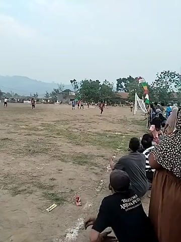 A group of people are watching a soccer game on a dirt field. The spectators are sitting and standing along the edge of the field, some are wearing headscarves and casual clothing. There are players on the field in the distance, wearing jerseys and shorts, and they appear to be playing soccer. There is a soccer goal on the right side of the frame with a flag above it. The background shows trees and houses, with hills in the far distance. The sky is overcast.