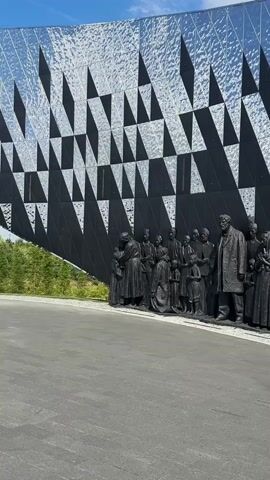 The video shows a row of dark, life-sized bronze statues depicting people of various ages, from adults to children, standing in a line against a black building with a reflective, geometric facade. The statues are rendered in a realistic style, with many of them appearing to be in poses of contemplation, grief, or quiet waiting. Some are looking down, others are embracing, and some are holding small objects. In the foreground, a young woman wearing a plaid shirt and shorts is visible, facing away from the camera and towards the statues, appearing to take a picture or observe them. The ground in front of the statues is paved with asphalt and a textured border of gravel. The lighting suggests it is daytime, with sunlight reflecting off the building's facade and casting shadows. The overall impression is one of solemnity and historical remembrance.