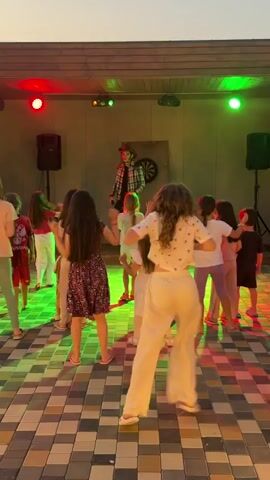 A group of children are dancing in front of a man on a stage. The man is wearing a plaid shirt and a hat and appears to be singing into a microphone. The children are also dancing and singing along. The stage is lit with colored lights, and there are speakers on either side of the stage. The floor is tiled, and the walls are a neutral color. There is a dartboard on the back wall of the stage.