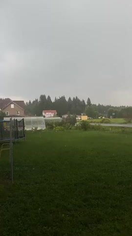 The video shows a backyard with green grass and a trampoline on the left. In the background, there are houses, trees, and a road. The sky is overcast and appears to be raining. Small white dots, likely raindrops, are visible falling in the air.