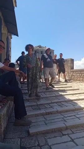 The video shows a group of people on a stone stairway outdoors under a clear blue sky. In the foreground, a person is seated on the left, wearing a dark shirt and pants. Moving up the stairs, a woman in a patterned dress is walking. Behind her, a man in a dark t-shirt and shorts, wearing a hat, is also walking up the stairs. Further up the stairs, other individuals are visible, including a woman in a blue dress and another person in a white shirt and light-colored pants. The environment appears to be a historic or tourist location, with stone buildings and pathways. The lighting suggests it is daytime, likely sunny.