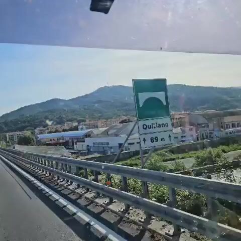 The video is shot from a moving vehicle, likely a train or bus, traveling on a bridge. The camera is pointed towards a town nestled at the base of a mountain. The town consists of a mix of residential and commercial buildings, and a church steeple is visible. A large industrial building with a light blue roof and a sign that reads "APERURA NUOVI REPARTI" is prominently featured in the foreground. The mountain is covered in lush green vegetation. In the distance, two wind turbines can be seen on the mountain's peak. The sky is blue with some clouds. A metal railing runs along the side of the bridge. The video is slightly shaky due to the vehicle's movement.
