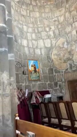 The camera films from a low angle, showing a small group of women singing in a church. The women, dressed in burgundy robes and white head coverings, are standing near a music stand. They appear to be singing from sheet music held in their hands. They are in a domed room made of gray stone. A framed painting hangs on the wall behind them. There are several rows of wooden pews in front of them.