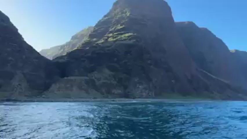The video shows a seascape with tall, rocky cliffs covered in greenery. The water is turquoise and choppy, with small waves breaking on the shore. The sky is clear and blue. There is some sound that includes music and speech.