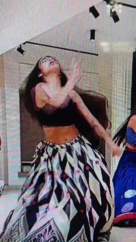 A young woman with long black hair dances in a room. She is wearing a black crop top and a long skirt with a black, white, and red diamond pattern. Her arms are extended and her body is in motion. The room appears to be a studio or performance space with spotlights on the ceiling and a window with blinds in the background.