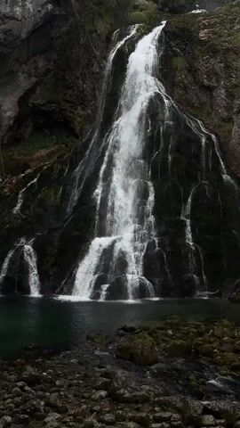 The video shows a tall waterfall cascading down a rocky cliff face into a pool of dark green water. The cliff is covered in moss and vegetation, and the water creates a misty spray as it falls. The surrounding area is rocky and covered in loose stones.