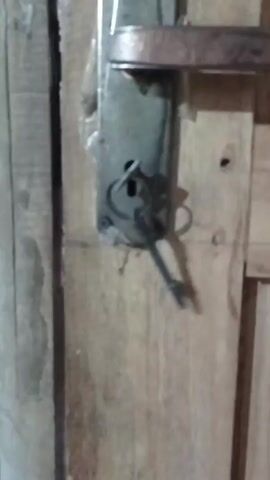 The video shows a close-up of a door with a lock and key. A metal key is being inserted into the lock. The door appears to be made of wood, with visible grain and texture. The lock mechanism is also metal and shows signs of wear. The lighting is dim, creating shadows on the door and lock.
