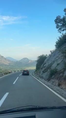 A car drives on a mountain road with a view of a valley and the sea. The sky is blue with a few clouds. The road is paved and has white lines. The car is black and has its brake lights on. There are trees and rocks on the side of the road.