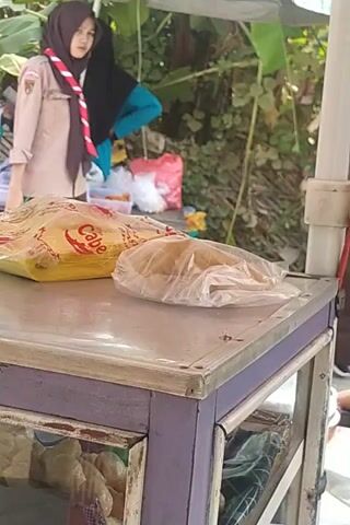 A young person in a scout uniform, including a headscarf, is standing next to a mobile food cart. Another person is seated behind the cart. The food cart contains packaged food items. The scene appears to be outdoors, with greenery in the background. The camera angle is tilted.