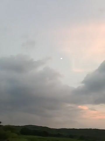 The video shows a slightly blurry daytime outdoor scene with a vast sky filled with clouds and a faint light source, possibly the sun. The lower portion of the video reveals a dark, hilly landscape with some sparse vegetation and a hint of greenery in the foreground. The sky transitions from a pale, hazy white and pink near the horizon to a lighter gray and white higher up. There are no people or objects clearly visible in the video.