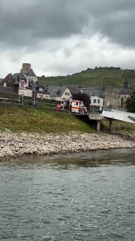 The video shows a river with choppy grey water in the foreground. In the background, a small town is nestled on a hillside covered in vineyards. A white bridge arches over the river towards the town. On the riverbank, near the bridge, an ambulance with flashing lights is parked, along with several people dressed in red and white uniforms, some of whom appear to be emergency responders. They are gathered around the ambulance and near the bridge, suggesting a possible rescue or medical incident. The sky is overcast with dark clouds, and the overall atmosphere is somber.