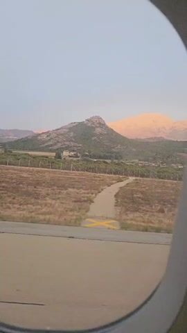 The video shows a view from an airplane window. In the foreground, a section of an airport runway is visible. Beyond the runway, there is a field of dry, brown grass, bordered by a fence. In the background, a prominent, rocky mountain covered in green vegetation rises against a pale sky. The peaks of more distant mountains are illuminated with a warm, rosy light, suggesting it may be sunrise or sunset. A dirt path winds through the grassy field. There are no people or animals visible in the frame. The camera is stationary, and the view is consistent throughout the short clip.