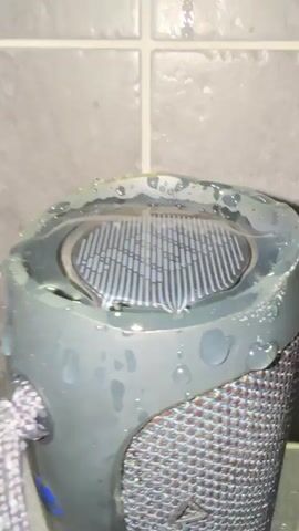 A close-up shot of a black speaker with a grey and white rope handle attached. The speaker is wet, with water droplets visible on its surface and pooled on the top. The background consists of grey tiled walls. The speaker is placed on a surface, possibly a counter or shelf. The video shows the speaker with water on it and in the immediate vicinity.
