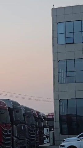 A line of parked semi-trucks is shown with a building to the right. The sky is a light orange hue, suggesting it is either sunrise or sunset. The trucks are all facing left and appear to be new or well-maintained. The building is modern with large windows. There is no discernible action or interaction occurring in the video.