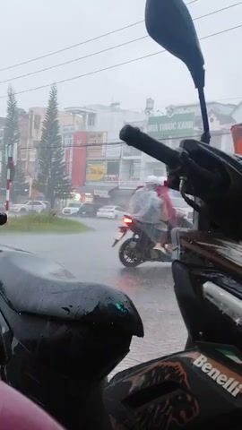 A person on a motorcycle wearing a light-colored rain poncho rides through heavy rain on a street lined with buildings and trees. The motorcycle in the foreground is dark-colored and wet. Other vehicles, including cars, are visible on the road, also driving through the rain. The sky is overcast and the weather is poor.