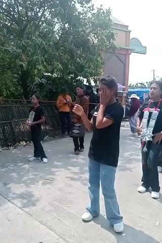 A group of young women are dancing outdoors. The woman in the foreground is wearing a black t-shirt and blue jeans, and she is dancing with her arms outstretched. Behind her, other women in black t-shirts are also dancing. In the background, there are trees, buildings, and other people watching. The ground is paved with concrete. The overall atmosphere appears to be festive.