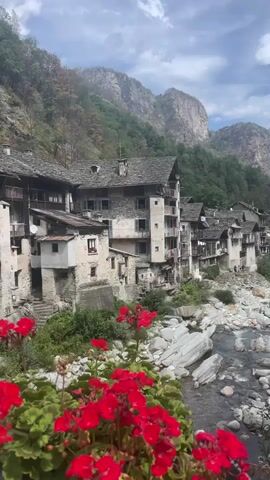 The video pans across a picturesque village nestled in a mountain valley. The architecture is rustic, with stone buildings featuring wooden balconies and slate roofs, suggesting an older, traditional settlement. A clear stream with smooth rocks flows in the foreground, and vibrant red flowers are in bloom, adding a splash of color to the scene. The background is dominated by steep, rocky mountains covered in lush green vegetation, with a partly cloudy sky overhead. There are no people or visible actions in the video, creating a serene and tranquil atmosphere.