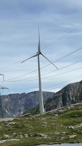This media file captures a scenic mountain landscape dominated by a tall, white wind turbine. The turbine's three blades are visible, two of which are angled upwards and to the side, while the third points downwards. The tips of the blades are marked with red. The turbine stands on a rocky, grassy hillside dotted with boulders. In the background, rugged, grey mountains rise steeply, with patches of snow visible on their peaks, suggesting a high-altitude location. Power lines crisscross the frame, with a large electrical pylon visible on the left.

The sky is a bright blue with scattered white clouds, indicating fair weather and likely daytime. A small body of water, possibly a pond or lake, is present in the foreground.

A prominent road sign is visible in the middle of the frame, with text in Italian. The top sign, blue with white text, reads "TRANSITO" with an upward-pointing arrow. Below it, a white sign with black text says "Ospizio" and features symbols for parking (a 'P'), a bed, and crossed-out cutlery, indicating accommodation and possibly a restaurant. The bottom sign, brown with white text, reads "Museo/Sasso San Gottardo" with a right-pointing arrow. This text provides significant location cues, pointing towards the Gotthard Pass region in Switzerland, famous for its transit routes, hospice (Ospizio), and the Sasso San Gottardo Museum.

The overall impression is one of a serene, remote, and perhaps windswept mountain environment where renewable energy infrastructure is integrated into the natural landscape.