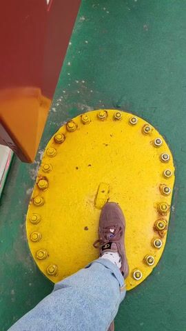 A first-person perspective shows a person standing on a yellow, oval-shaped hatch cover, rimmed with bolts, on a green surface. The person is wearing a brown sneaker, socks with a pattern, and blue jeans. A red and yellow structure is visible to the left of the hatch.