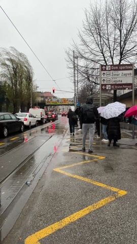 The video depicts a street scene on a rainy day. Several people stand under umbrellas at a tram stop, waiting. Cars are stopped at a red light on the wet street alongside the tram tracks. A sign indicates directions to various locations including Dom sportova, Zoonar Zagreb, Laguna, and KC Drazen Petrović. The sky is overcast, and the trees are bare, suggesting late autumn or early spring. The ground is wet and reflections of the red traffic lights can be seen on the pavement. Some people are walking by, holding umbrellas. Yellow zig-zag lines are painted on the sidewalk near the tram stop.