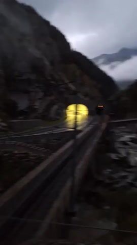 A blurry, dark video captures a winding road in a mountainous environment. The road appears to be paved and has guardrails. There are tunnels visible in the mountainside, with one tunnel illuminated by a bright yellow light. Several vehicles are seen on the road, including a blue car and a white car. The weather appears overcast and foggy, with clouds obscuring the tops of the mountains. In the foreground, there is a fence and some vegetation. The video is shaky, suggesting it was filmed from a moving vehicle.