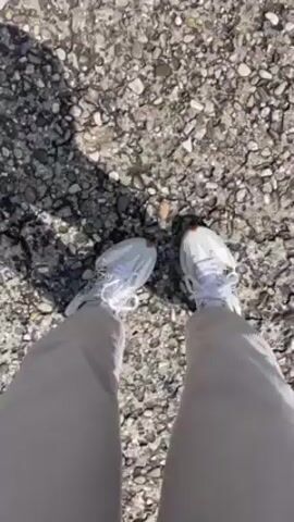 A person wearing white sneakers and light beige corduroy pants stands on a surface covered in small gray and white rocks. The camera, positioned above, shows the person's legs and feet as they shuffle slightly on the rocky ground. The sun is out, casting shadows across the gravel.