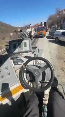 The video presents a first-person view from the operator's seat of a road milling machine. The machine is moving along a paved road, and in the background, other vehicles, including a truck and a pickup, are parked alongside the road. A second milling machine is visible ahead, working on the road surface. The dashboard of the machine shows digital displays with various readings. Outside the machine, trees line the side of the road and buildings are visible on a hillside. The sky is clear and blue.  The operator is wearing black trousers and shoes, and is sitting behind the steering wheel.