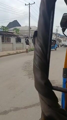 The video shows a view from inside a yellow and blue auto rickshaw moving down a street. A thick, gray, leather-like material is wrapped around the support column of the rickshaw, partially obstructing the view. The street is paved and shows some wear and tear. In the background, there are various vehicles, including a blue sedan, a white pickup truck, and another blue auto rickshaw. There are buildings on either side of the street, some residential and others commercial. The sky is overcast and a person wearing a red shirt can be seen walking on the left side of the street. The rickshaw is in motion.