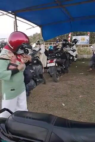 The video shows an outdoor scene, possibly a parking area, under a blue tarp awning. Several motorcycles are parked in a row. In the foreground, a person wearing a light green shirt, white pants, a red helmet, and a mask is standing and interacting with something in their hands. A motorcycle is partially visible in the bottom foreground. In the background, other individuals are seen on or near motorcycles, and there are some structures and vegetation. One person in the background is wearing a light-colored shirt and a helmet, sitting on a motorcycle. The ground appears to be dirt and grass. The lighting suggests it is daytime.