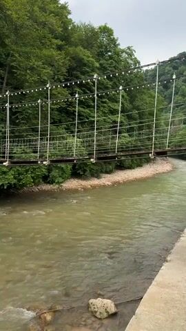 The video shows a view of a suspension bridge over a river. The bridge is made of wood and metal with cables supporting it. The river is flowing with murky water and rocks visible on the riverbed. The banks of the river are lined with lush green trees and foliage. The overall scene depicts a natural landscape with a man-made structure.