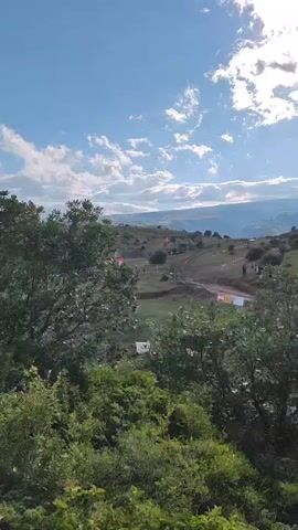 The video captures an outdoor scene with rolling hills and a bright, partly cloudy sky. In the foreground, lush green trees partially obscure the view. Further back, a dirt track winds through the grassy terrain. Several off-road vehicles, some appearing to be ATVs or similar recreational vehicles, are visible on and around the track. Red flags are planted along the course, marking the path or indicating event locations. A few people are seen standing or walking near the track and the vehicles. The overall impression is of an off-road racing or recreational event taking place in a natural, hilly landscape.