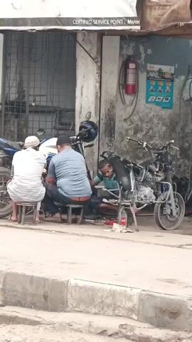 Three men are working on a motorcycle in front of a motorcycle repair shop. Two men are seated on stools, while the third man is crouched down working on the motorcycle's engine. Another motorcycle in a state of disassembly is on a stand nearby. The shop's sign reads "CERTIFIED SERVICE POINT : Moto". A fire extinguisher hangs on the wall. The environment is urban, with a concrete sidewalk and street.