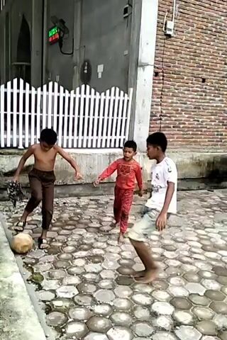 Three barefoot boys are playing with a coconut in an outdoor area. The boy on the left is shirtless with brown pants, and the boy in the middle wears a white t-shirt with the number 9 on the back and denim shorts. The third boy wears a dark t-shirt and denim shorts. They are playing with the coconut on a wet, paved area with a hexagonal pattern. Behind them is a building with a brick wall, a white picket fence, and a doorway. A small digital clock is mounted on the wall.
