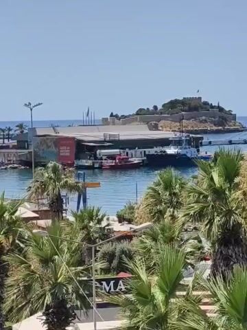 This video captures a bright, sunny day at a bustling harbor, likely in Kuşadası, Turkey. The scene is dominated by a large cruise ship, partially visible on the right side of the frame, docked near an island fortress. The fortress, with its stone walls and a flag flying, stands prominently in the background against a clear blue sky.

In the foreground, lush palm trees sway gently, framing the view of the harbor. Between the palm trees and the water, there are several boats docked, including a red vessel and a blue and white tugboat. The water is calm and reflects the bright sunlight. A building with signs is visible on the left, with some text partially obscured by palm fronds. One visible sign appears to say "GATES OF HISTORY."

The overall atmosphere is one of activity and vacation. The clear sky and bright sunlight suggest it is midday, and the presence of a cruise ship and harbor activity points to a popular tourist destination. There are no clearly visible people in the foreground, but the environment suggests a lively place. The video pans slowly, offering a panoramic view of the harbor and its surroundings.