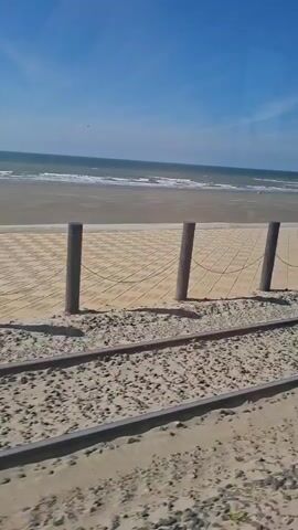 The video shows a beach with a wooden boardwalk and sand. In the foreground, there are two sets of railroad tracks running parallel to the beach. The boardwalk is elevated and has wooden posts with cables strung between them. The ocean is visible in the background with waves rolling onto the shore. The sky is clear and blue. The video appears to be taken from a moving vehicle, possibly a train, as indicated by the tracks and the slight blur of motion. There are no people visible in the video. The scene suggests a sunny day at the beach, possibly a tourist area with a unique transportation system.
