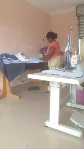 A woman in an orange shirt and yellow and white patterned pants stands at a sewing table, working on a light blue garment. She is facing away from the camera. To her left is a larger, dark blue table with more light blue garments on it. A sewing machine is visible to the right of the woman, with a pink basket and other items nearby. The room has pale pink walls and a tiled floor.