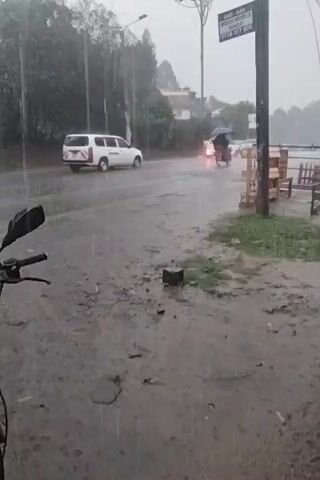 The video shows a street during a heavy rain. A white van and a small motorbike with a black umbrella are driving on the road. The road is wet and there are puddles. On the side of the road, there's a sign that reads "WOOD WORKS SHOP," and some wooden benches. The ground is muddy. The video appears to be taken from a stationary position. The weather is overcast.