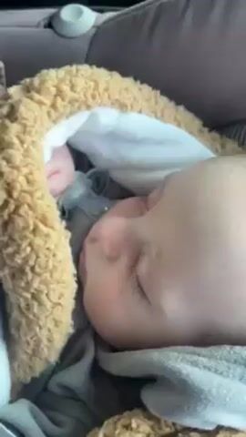 A baby is sleeping in a car seat. The baby has a pacifier in their mouth and is secured in the seat with a safety belt. The baby is wearing a grey sweater and has a light brown plush support around their head. The surrounding interior of the car is visible, including the car seat and a portion of the dashboard.