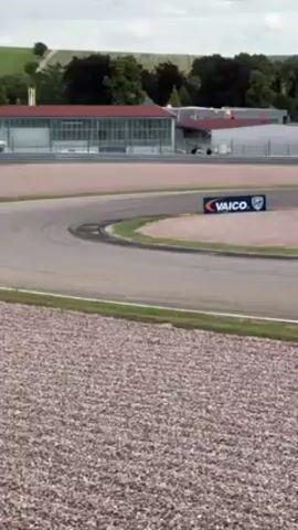 The video shows a white Porsche race car driving on a racetrack. The car is moving from right to left, negotiating a curve in the track. The environment includes a gravel section in the foreground, the racetrack, and buildings and trees in the background. There is also a Vaico sign along the side of the racetrack. The lighting suggests it is daytime with overcast conditions.