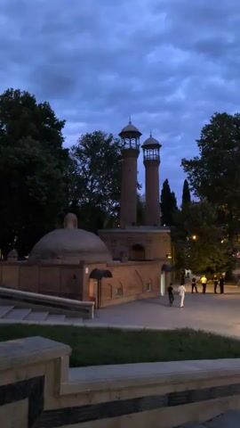 The video captures a dusk scene in what appears to be a historical area. In the foreground, a stone structure with a patterned border is visible, suggesting it's part of a pathway or staircase. The mid-ground features a prominent brick building with a domed roof on the left and two tall, slender towers, reminiscent of minarets, on the right. The architecture suggests a mosque or a similar religious structure. Trees with dense foliage flank the building and surround the area, with some trees in the foreground and others behind the main structure. A lamppost with an ornate fixture illuminates a section of the paved pathway in the mid-ground. Several individuals are present in the scene. On the pathway, a few people are walking, including one person in white attire. In the distance, near the building, a small group of people are gathered. The sky above is a deep blue, indicative of twilight, with visible clouds creating a dramatic effect. The overall atmosphere is serene, with the lighting suggesting the transition from day to night.