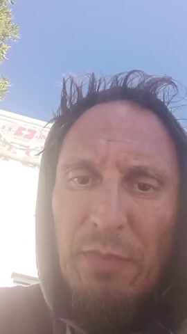 The video shows a man wearing a hoodie. He is looking directly into the camera and talking. Behind him is a blue sky and part of a building with a sign on it. The sign is partially visible but not readable. The man has short, dark hair that is slightly messy. He has stubble on his chin and a gap between his front teeth.