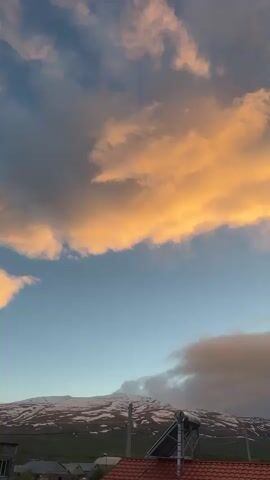 The video displays a time-lapse of clouds in a partly cloudy sky during dusk. The clouds are colored in hues of orange and pink, suggesting the sun is setting or has recently set. In the foreground, the rooftops of houses and a solar panel can be seen. In the background, there is a mountain range with some snow visible on the peaks. The sky gradually changes from deeper blues to lighter pinks as time progresses, and the cloud formations also shift and dissipate over time. Throughout the time-lapse, the wind is blowing audibly.