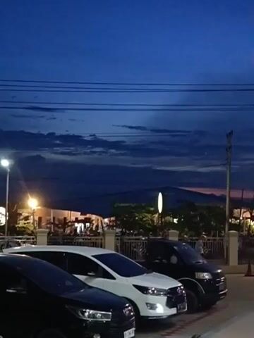 The video shows a view of parked cars against a twilight sky. The sky is a deep blue with streaks of darker clouds. There are power lines running horizontally across the upper part of the frame. In the middle ground, there are streetlights and buildings illuminated with warm light. The foreground is dominated by the tops of several cars, including a black car on the left, a white SUV in the center, and a black van on the right. The cars are parked in a lot. The overall atmosphere is calm and dusky.