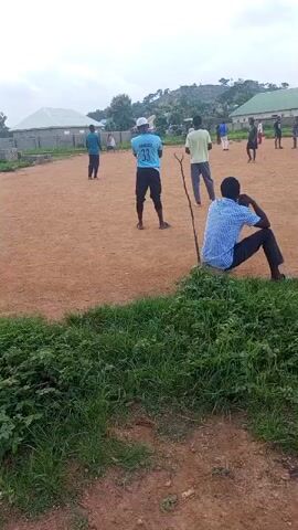 The media file shows an outdoor scene in Kagarko South, Nigeria. The foreground is dominated by lush green grass and vegetation, suggesting an area that is not heavily manicured. In the background, a sandy or dirt field is visible, where a group of people are gathered. The field appears to be a gathering space, possibly for recreation.

Several individuals are scattered across the field. In the middle ground, a person wearing a blue t-shirt with the number "33" prominently displayed on the back stands facing away from the camera. They are also wearing a white cap and dark pants. To their right, another person in a light yellow t-shirt and grey pants is also standing. Further in the background, more people are visible, some appearing to be in motion, suggesting an activity or game is in progress. One person in the foreground, to the right of the frame, is seated and observing the scene. They are wearing a blue and white checkered shirt and dark pants. A thin stick is planted in the ground near them.

The setting includes buildings with light-colored roofs in the distance, hinting at a residential or community area. Hills or elevated terrain are also visible in the far background. The sky is overcast, suggesting a cloudy or potentially impending weather event, though the lighting does not indicate harsh sunlight, implying it might be morning or late afternoon. The overall atmosphere appears relaxed and informal, with people engaged in some form of communal gathering or activity. The visible text on the t-shirt reads "DAKAR", "33", and potentially another word above it.