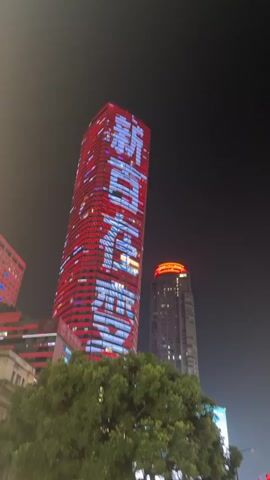 The video displays a nighttime urban landscape featuring several illuminated skyscrapers. One prominent building on the left is tall and rectangular, its facade adorned with red and white lights that appear to form text or patterns. To its right, another skyscraper is visible, also with red lighting. In the center, a shorter, rectangular building has a golden illuminated sign. A third, cylindrical skyscraper on the right is covered in bright red lights, creating a striking visual effect against the dark sky. The lower part of the frame shows the tops of green trees, suggesting the viewpoint is from ground level or a low-rise structure. The overall atmosphere is one of a modern, brightly lit city at night.