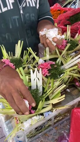 The video shows a person’s hands arranging flower arrangements in a container. The arrangements are small, circular, and made of woven leaves. They contain pink and white flowers, as well as some green foliage. The person is wearing a green shirt and a red and white bracelet. The arrangements are in a metal basket. There are red plastic bags in the background. The person is adding white flowers to the arrangements.