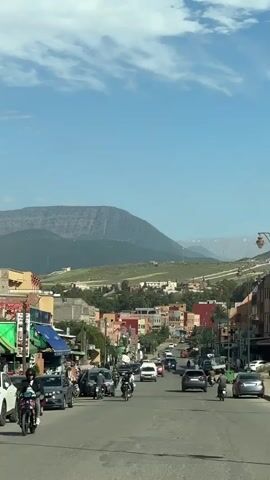 The video shows a street in a town with mountains in the background. There are cars and motorcycles on the street, and people walking on the sidewalks. The buildings are a mix of colors, and there are trees lining the street. The sky is blue with some white clouds. The camera appears to be in a car moving slowly down the street. Music is audible.