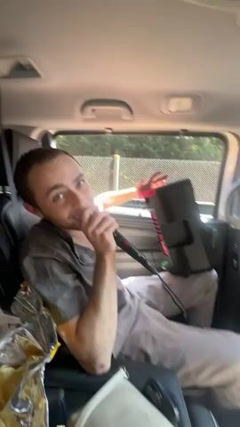 A young man with short brown hair, wearing a grey t-shirt and light-colored pants, is seated in the backseat of a car during the daytime. He is holding a black microphone with a red band and singing or speaking into it, looking towards the camera with an engaging expression. A black portable speaker rests near his lap. On the seat beside him, there is a silver foil-like bag, possibly containing food or snacks. A car seatbelt is visible to his left. Through the car window, sunlight streams in, and a mesh fence with greenery can be seen in the background. Another person's hand, holding a red object, is briefly visible through the window, appearing to interact with the outside. The man points his finger at one point during his performance. A small abrasion or wound is visible on his right elbow. The scene suggests a casual karaoke or performance session inside the car. The location is Coajomulco, Mexico.