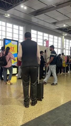 The video shows an indoor scene, likely an airport or train station, with several people in line. In the foreground, a man wearing a dark vest over a light-colored shirt and dark pants stands with a black suitcase. To his left, a woman in a pink shirt and jeans stands with a black backpack. Behind them, a group of people are queuing. The queue is organized with stanchions. A digital display board is visible on the left, showing an advertisement with yellow and blue colors and text. The environment appears to be a well-lit space with large windows and a tiled floor. The people in the line are diverse in age and gender, and most are carrying bags or luggage. Some individuals are looking at their phones or talking to each other while waiting. The overall atmosphere suggests a public transit hub during a busy period.